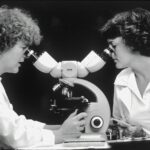 Black and white photo of two female-presenting scientists looking into adjoining microscopes